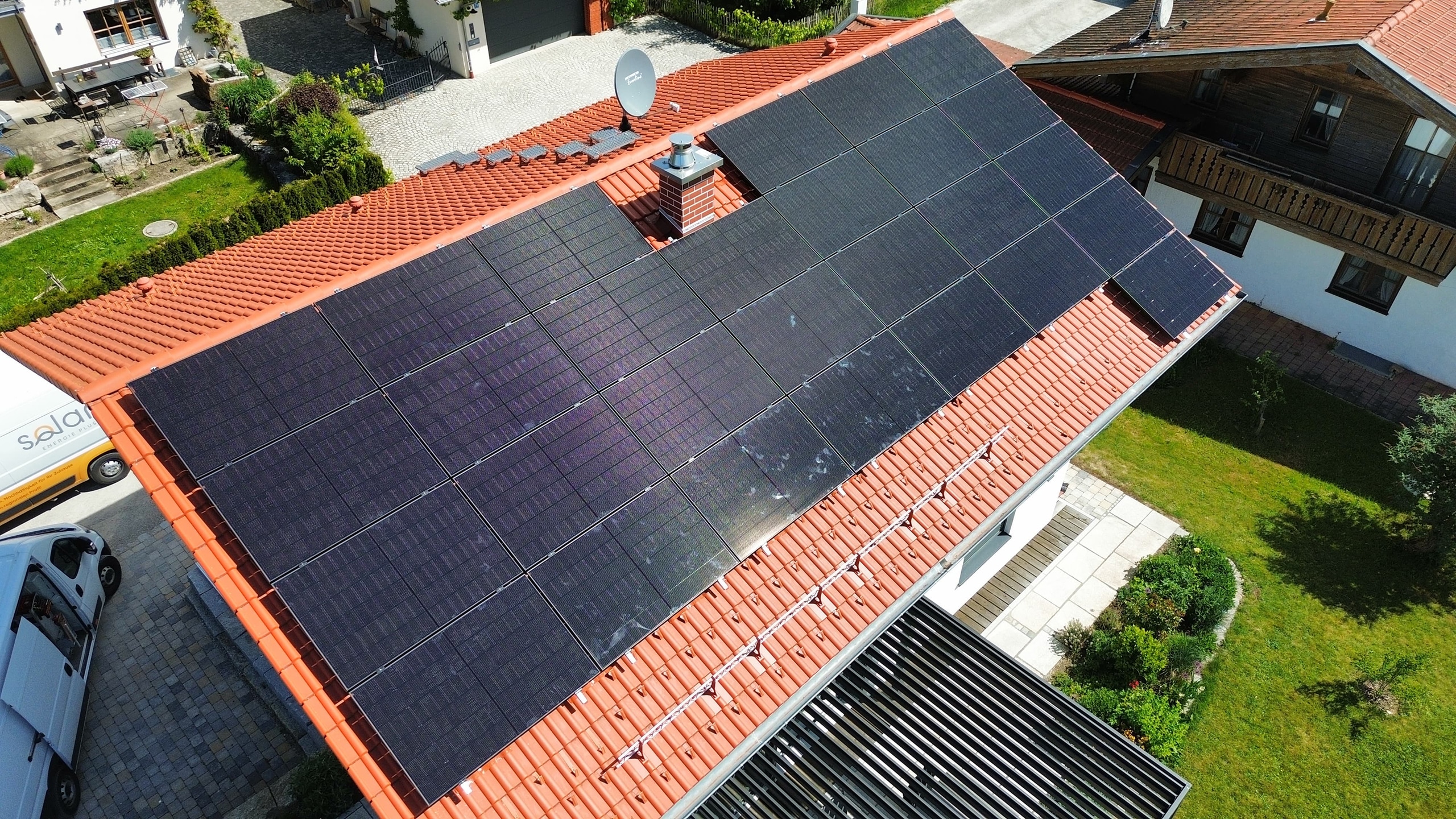 Solaranlage_SolarenergiePLUS Eine Photovoltaikanlage auf einem roten Ziegeldach in vollflächiger Belegung. Das Haus steht in einem gepflegten Garten mit begrünter Fläche und gepflastertem Eingang.