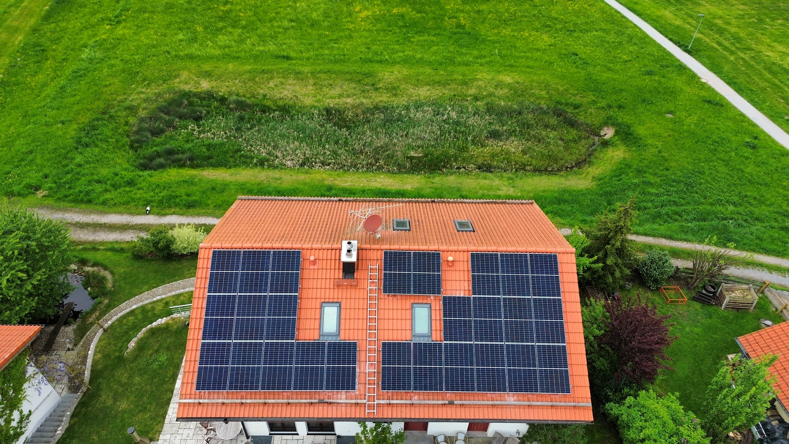 Solaranlage-Privat_SolarenergiePLUS Eine private Solaranlage auf einem roten Ziegeldach. Das Haus steht inmitten eines großen grünen Grundstücks mit Wegen, Bäumen und Gartenflächen.