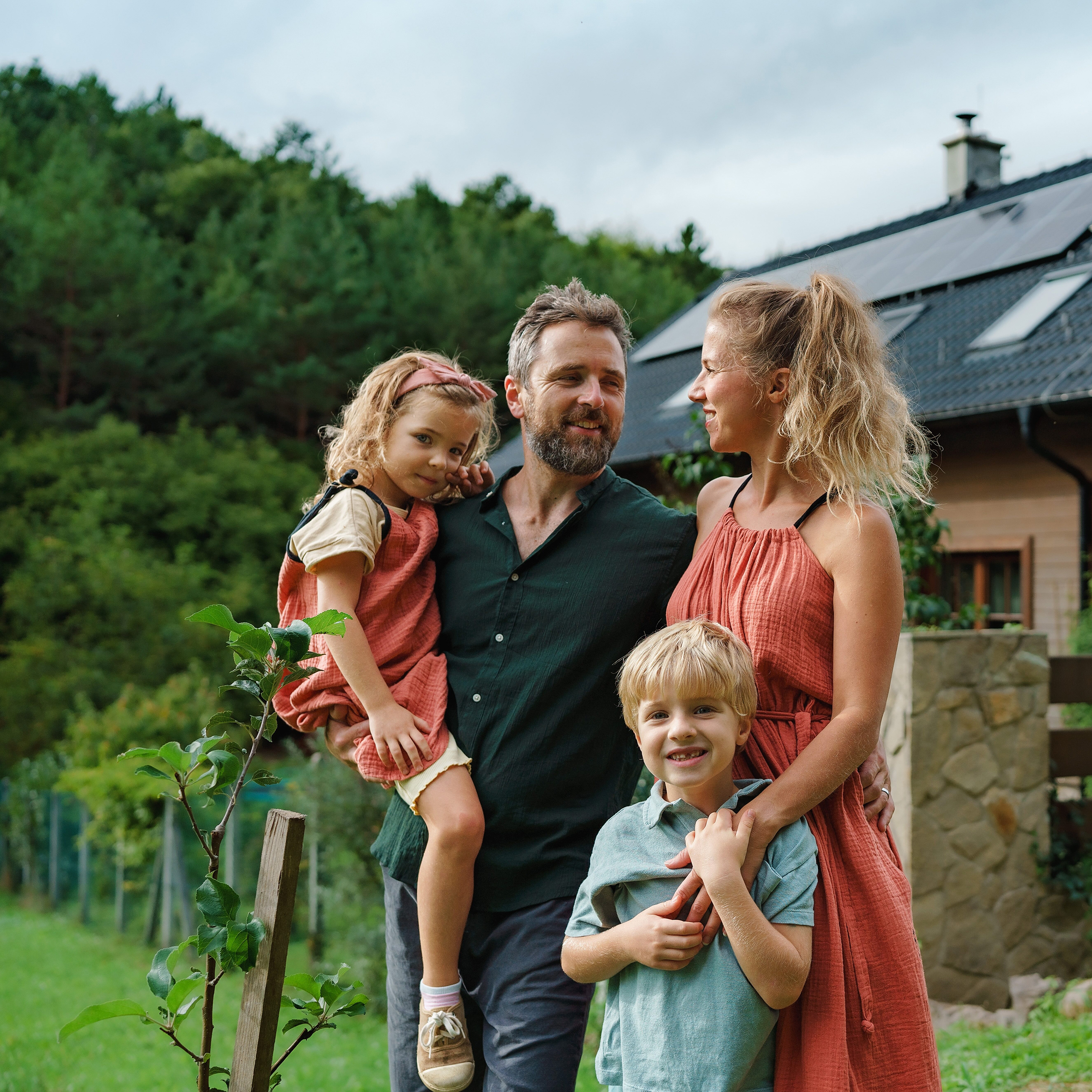 shutterstock_2206914219 Eine Familie, bestehend aus Mutter, Vater, Tochter und Sohn, im Garten vor einem Wohnhaus mit installiertem Solarsystem auf dem Dach. Die vier Personen stehen nah beieinander und wirken entspannt im grünen Umfeld.