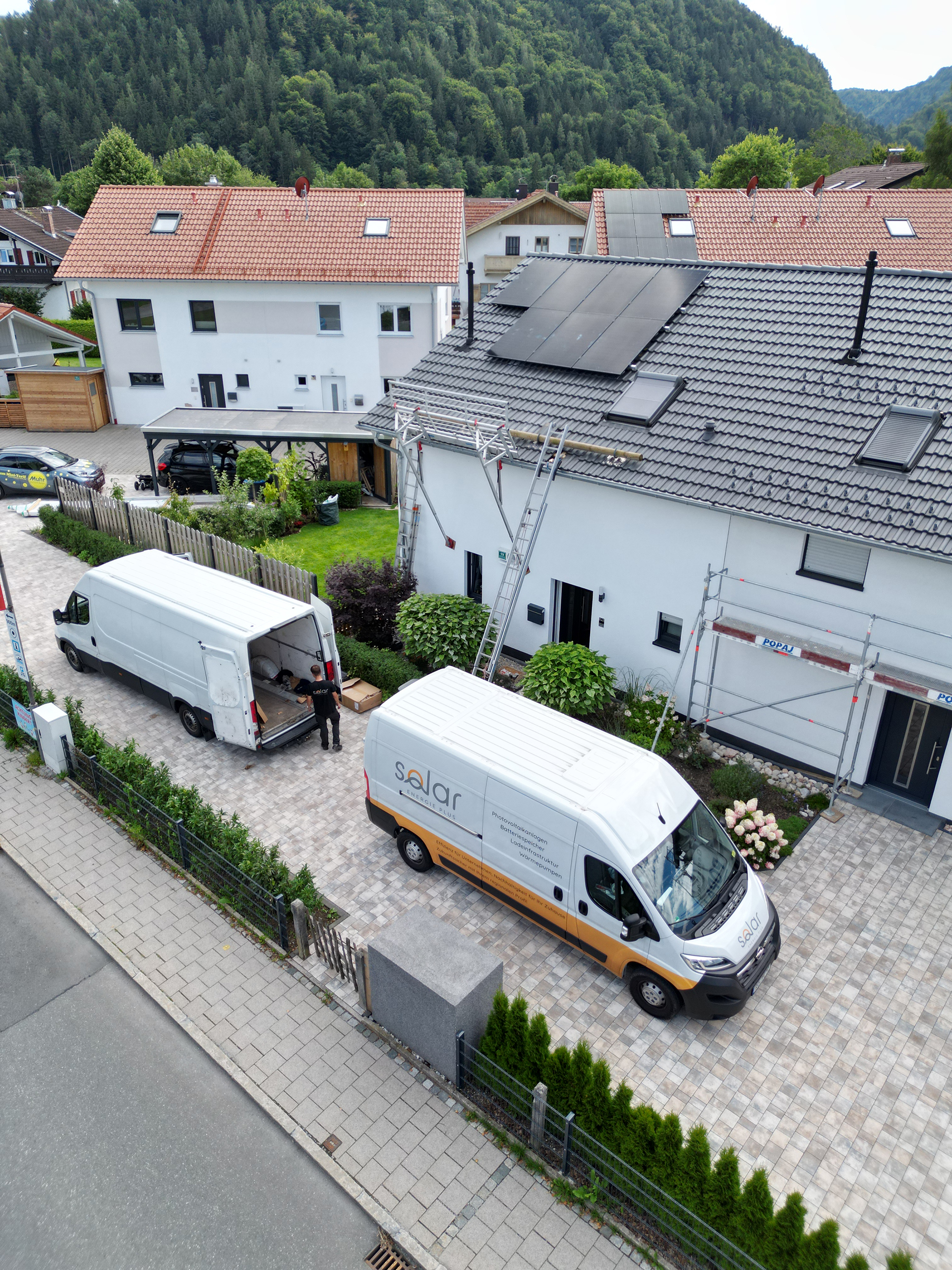 Privatkunden_Batteriespeichersysteme_2 Eine Photovoltaikanlage auf dem Dach eines Wohnhauses in einer Wohnsiedlung. Servicefahrzeuge für Privatkunden-Batteriespeichersysteme stehen in der Einfahrt.