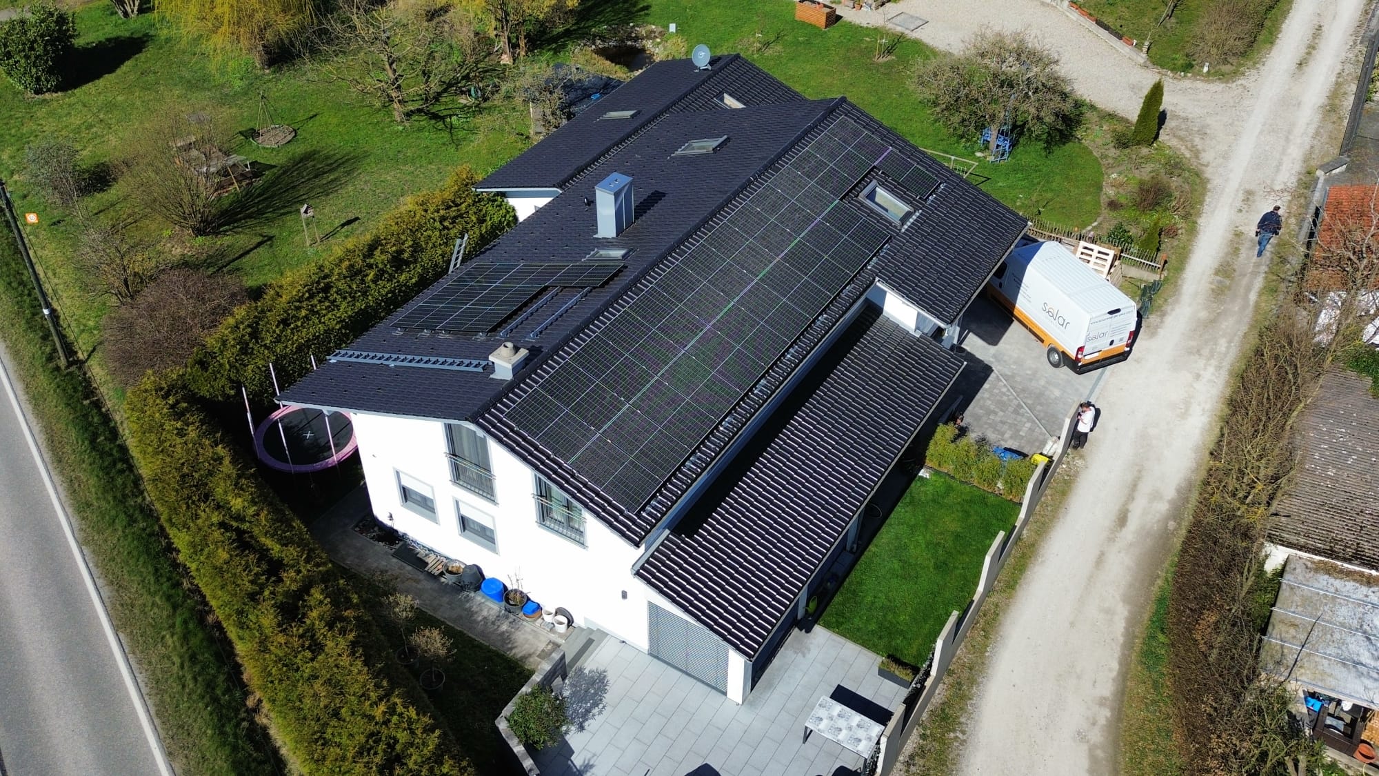 Privatkunden_Photovoltaik_1 Eine große Photovoltaikanlage auf dem Dach eines modernen Einfamilienhauses. Das Grundstück ist weitläufig und von Grünflächen umgeben, während ein Servicefahrzeug in der Einfahrt steht.
